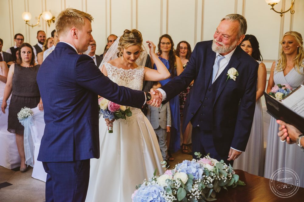 The groom shakes the hand of his father-in-law as he escorts his daughter down the aisle at the wedding ceremony in Gosfield Hall's Grand Salon