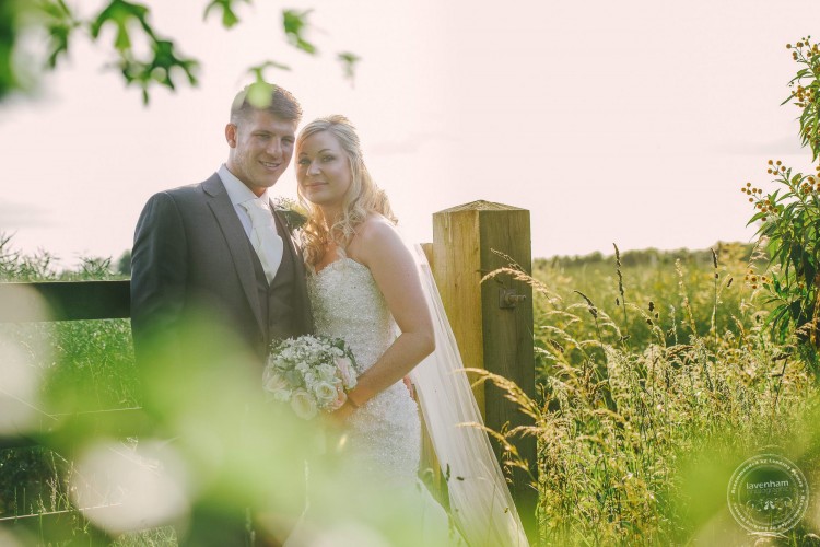 Bride and groom at gate with field and crops, Crabbs Barn. Wedding Photography by Lavenham Photographic