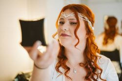 A bride with fiery red hair styled in loose curls wearing a delicate tiara reflects on her wedding preparations, showcasing her elegant jewelry and radiant look before her special day. Elegant red-haired bride with a silver tiara and pendant necklace, capturing her reflection in a mirror, preparing for her wedding at a venue in Essex or Suffolk.