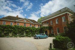 Elegant wedding transportation at a historic Essex or Suffolk venue, capturing timeless charm and perfect photo opportunities. Vintage blue car parked in front of a classic red brick estate with lush greenery and a bright blue sky.