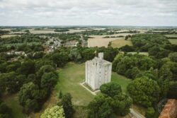 Aerial view of a historic castle tower amid dense greenery with rural fields and a small village in the background. Ancient castle tower surrounded by lush green trees in a scenic countryside landscape of Essex and Suffolk.