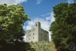 Historic castle tower framed by vibrant green foliage under a blue sky, ideal for wedding photography in Essex and Suffolk. Ancient castle tower surrounded by lush green trees against a bright blue sky with scattered clouds in Essex and Suffolk.