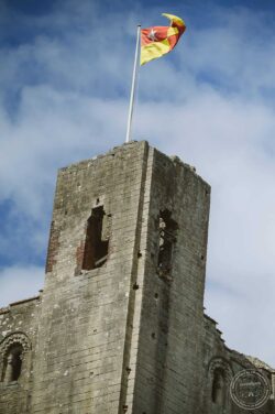 Historic castle tower with waving flag, captured in Essex & Suffolk, highlighting heritage and scenic views for weddings and photography. Rustic stone castle tower with a windswept flag flying at the top, set against a bright blue sky with clouds, showcasing historical architecture in Essex & Suffolk.