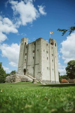 A historic stone castle with tall towers and a flag, set against a vibrant blue sky, surrounded by lush greenery, perfect for capturing timeless wedding memories in Essex or Suffolk. Medieval castle with stone walls, tall towers, and a flag flying under a bright blue sky with scattered clouds, ideal for romantic wedding photography in Essex & Suffolk.