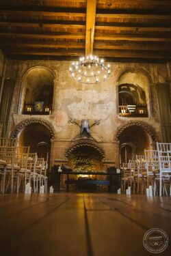 Elegant rustic wedding interior with medieval armour, candle decorations, wooden chairs, and a grand fireplace, creating a romantic and historic atmosphere for wedding photography in Essex and Suffolk. Stone fireplace with medieval armor and crossed swords, surrounded by candlelit decorations and rustic seating, perfect for a historic wedding ceremony at a vintage Essex and Suffolk venue.