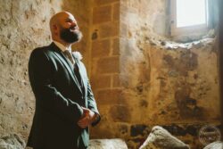 A groom in a suit and tie stands joyfully in a rustic stone church, illuminated by natural light, ready for his wedding vows. Bald groom with beard in tuxedo inside an ancient stone chapel with sunlight streaming through a small window, capturing a special moment before his wedding.