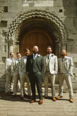 A group of stylish wedding groomsmen in beige suits and floral boutonnieres in front of a historic stone church in Essex & Suffolk, shot by Lavenham Photographic. Elegant wedding groomsmen standing outside historic church entrance in Essex & Suffolk, dressed in beige suits with boutonnières, captured by Lavenham Photographic.