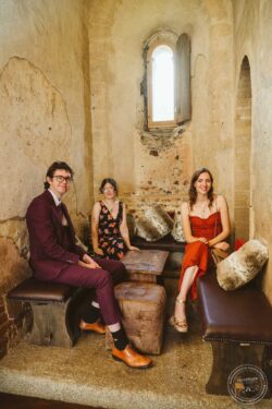 Group of three friends smiling and sitting inside a historic, rustic building with stone walls and arched window, dressed stylishly for a wedding day. Vintage Indoor Portrait of Three Friends Relaxing in Rustic Wedding Venue, Essex & Suffolk.