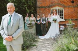 A joyful wedding celebration captured outdoors with the bride, bridesmaids, and groomsmen in front of a historic brick house, surrounded by lavender and decorative planters, showcasing wedding attire and happy moments. Elegant bride with red hair in a flowing white gown holding a bouquet, standing alongside bridesmaids and groomsmen in formal attire during a wedding photoshoot outside a charming brick building.
