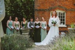A beautiful wedding scene captured outdoors, featuring the bride with her bridal party during a sunny day, with lush greenery, lavender plants, and classic architecture creating a timeless romantic atmosphere. Elegant bride with vibrant red hair holding a white floral bouquet, surrounded by bridesmaids and groomsmen in soft green dresses and dark suits, in a picturesque garden setting with historic brick building background.