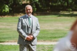 A joyful groom in a stylish grey suit with a green tie, standing outdoors on a bright day during a wedding ceremony, capturing a candid and emotional moment at a picturesque Essex or Suffolk wedding venue. Elegant grey-suit groom smiling outdoors on wedding day, lush green background, natural light, heartfelt moment, wedding photography Essex & Suffolk, Lavenham Photographic.