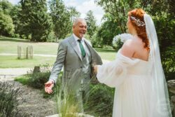 A bride with red hair in a flowing white wedding dress and veil celebrates a special day with her father in a scenic outdoor setting. Joyful wedding moment capturing a bride and her father sharing an emotional moment outdoors in a lush green garden in Essex and Suffolk.