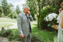 A touching moment at an outdoor wedding where a father in a light grey suit, emotional and teary-eyed, hugs his daughter in a wedding dress, in a scenic garden with trees and greenery. Tearful father in grey suit emotional wedding moment outdoors, bride holding bouquet of white flowers, lush green garden background, heartfelt father-daughter connection on wedding day.