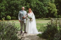 A joyful bride in a white wedding dress stands beside her father in a garden, holding a bouquet of white flowers under a bright sky with vibrant green trees in the background. Elegant bride with red hair in a white gown holding a bouquet, walking arm-in-arm with her father in a garden setting with lush green trees; wedding photography by Lavenham Photographic in Essex and Suffolk.