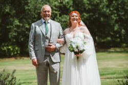 A joyful bride poses with her father during a wedding in Essex or Suffolk, wearing a beautiful off-the-shoulder wedding dress, holding a bouquet, with lush green trees behind them in a bright, natural setting. Elegant bride in a flowing white wedding gown holding a bouquet of white roses and greenery, smiling happily beside her proud father in a light grey suit outdoors on a sunny day.