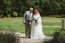 A joyful moment captured outdoors in the garden, showcasing the bride's elegant attire, her radiant smile, and the scenic natural background at a wedding venue in Essex or Suffolk. Beautiful bride with red hair and her father walking in a lush green garden on her wedding day. The bride holds a bouquet of white flowers, wearing a classic white wedding dress with off-shoulder sleeves and a floral headpiece.