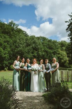 A joyful wedding group portrait of bridesmaids, bride, and groom outdoors in a beautiful garden setting with vibrant greenery and blue sky. Elegant wedding party standing outdoors in a lush green garden on a sunny day, capturing joyful memories of a wedding at Lavenham Photographic in Essex & Suffolk.