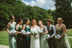 A joyful bride with her bridesmaids and groomsman sharing a happy moment outdoors at a wedding in Essex or Suffolk. Vibrant outdoor wedding photo capturing the bride and bridesmaids smiling and holding bouquets in a lush green garden setting.