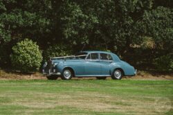 Elegant vintage blue Rolls-Royce automobile on a well-manicured lawn with a backdrop of dense greenery, perfect for capturing timeless wedding moments in Essex and Suffolk. Vintage blue classic car parked on a lush green lawn at a wedding in Essex & Suffolk, captured by Lavenham Photographic. Perfect for vintage wedding themes and nostalgic wedding photography.