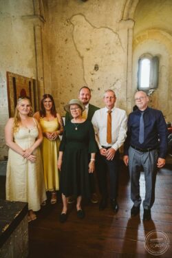 A group of six people, including family members, standing together inside a rustic, historic church with arched windows and textured stone walls during a wedding celebration in Essex and Suffolk. Family group portrait inside historic church at wedding reception Essex Suffolk wedding photographer.
