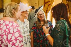A group of women dressed in colourful outfits and large elegant hats sharing a joyful moment during a wedding celebration in a rustic setting. Elegant women in floral and patterned dresses, wearing stylish wide-brim hats, enjoying a social gathering at a wedding reception in a warm, beautifully decorated venue in Essex & Suffolk.