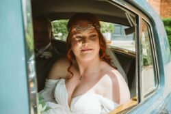 Bride seated in classic car, smiling and gazing out the window, showcasing her wedding dress and jewelry on her special day in Essex and Suffolk. Elegant bride with vibrant red hair looking through vintage car window on her wedding day in Essex, Suffolk, capturing a joyful moment before the ceremony.