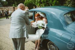 A bride with vibrant red hair in a beautiful white gown, holding a bouquet, as she exits a classic car on her wedding day. Elegant bride stepping out of a vintage blue car during her wedding in Essex, surrounded by greenery and guests.