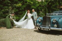 Bride and groom celebrating their wedding day with a vintage car at a lush outdoor venue in Essex and Suffolk. Elegant bride in a flowing white wedding dress and long veil with groom beside a vintage blue car, as bridesmaid adjusts the dress in lush garden setting, capturing special wedding day moments.