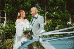 Elegant wedding couple standing together in a lush garden setting, with vintage car partly visible in the foreground, celebrating their special day. Beautiful bride and happy groom sharing a tender moment outdoors on their wedding day in Essex, captured by Lavenham Photographic.