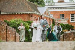 Beautiful wedding procession of bride, family and bridesmaids outside a historic brick building in Essex, captured by Lavenham Photographic. Elegant bride walking with her father and bridesmaids on a historic brick bridge during a wedding ceremony in Essex, captured by Lavenham Photographic.