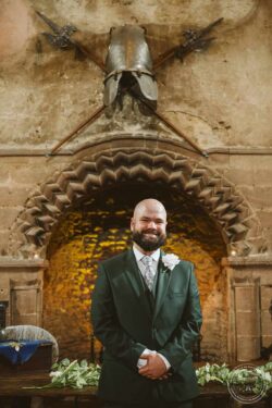 A portrait of a groom in formal attire at a wedding venue in Essex or Suffolk, featuring rustic stone architecture and medieval memorabilia, capturing a special moment on his wedding day. Elegant groom in a dark green suit with a white boutonniere, standing inside a historic stone building with medieval decor and a knight's helmet display above.