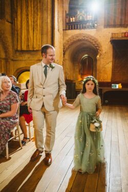 A groom in a beige suit and tie holding hands with a flower girl in a green dress with floral details, walking through the historic wedding ceremony space with seated guests and warm candlelight decor. Elegant groom holding flower girl’s hand at wedding ceremony in historic Essex venue, capturing joyful family moments, with warm lighting and rustic interior for timeless wedding photography.