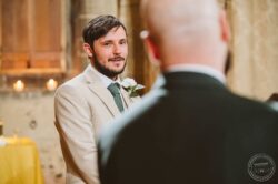 A heartfelt moment captured during a wedding at a historic Essex church, featuring a groom in a beige suit with a white rose boutonniere, celebrating love and commitment. Handsome groom in a beige suit with a white rose boutonniere gazing at the officiant during a wedding ceremony in an Essex church.