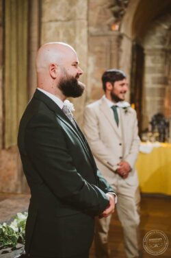 Joyful moment during wedding vows at a historic Essex & Suffolk venue, capturing the emotional connection between the grooms in a beautiful, timeless setting. Elegant gay wedding ceremony with two grooms in formal suits, standing indoors in a historic stone venue with warm lighting.
