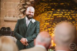 A happy groom in a tailored suit with a white flower boutonniere at his wedding in Essex, against a rustic brick wall, during a heartfelt moment at the ceremony. Bald groom with a beard in a dark green suit smiling during wedding ceremony at Essex & Suffolk venue.
