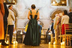 A beautifully captured moment of a wedding ceremony featuring a woman in a dark green satin dress with a lace-up back, set in a historic venue with candles and exposed brick walls, surrounded by well-dressed guests. Elegant woman in dark green satin dress with lace-up back during wedding ceremony at historic venue with exposed brick walls and flickering candles.