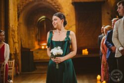 A bridesmaid in a beautiful emerald green gown smiles softly amidst seated guests in a warm, intimate wedding setting. Elegant wedding photograph capturing a bridesmaid holding a white rose bouquet during a ceremony in a historic, candle-lit venue in Essex & Suffolk.