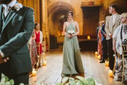 A joyful bridesmaid standing with guests seated along the aisle decorated with candles and petals in a charming, historic setting. Elegant maid of honour in a sage green dress holding white roses during a wedding ceremony in a rustic Essex & Suffolk venue.