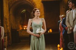 A bridesmaid in a pastel green dress stands gracefully holding a floral bouquet during a wedding in a rustic chapel with candlelit atmosphere. Elegant bridesmaid holding a bouquet of white roses during a wedding ceremony in a historic stone venue with warm candlelight ambiance in Essex & Suffolk.