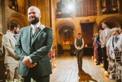 A heartfelt wedding moment captured inside an ancient church, with the groom waiting as guests watch and a bridesmaid holding flowers in the background; warm candlelight enhances the romantic atmosphere. Elegant groom in a grey suit and boutonniere waiting at an Essex and Suffolk wedding ceremony in a historic church, surrounded by attentive guests and warm candlelight ambiance.