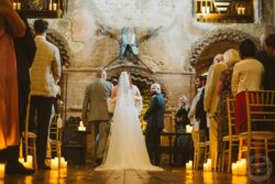 An intimate wedding ceremony taking place inside a medieval-inspired stone hall with soft candlelight, where the bride and groom exchange vows while guests witness the heartfelt event. Elegant wedding ceremony in a historic stone venue with romantic candles and medieval decor, featuring the couple exchanging vows surrounded by guests on a warm, rustic evening.