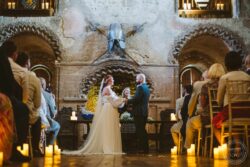 A romantic wedding in an ancient stone hall, capturing the bride and groom during their vows with guests seated around, candles creating a warm glow, and medieval decor including a mounted sword and shield. Elegant wedding ceremony inside a historic stone building with a medieval atmosphere, featuring a bride and groom exchanging vows amidst candles and guests.