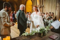 A beautiful wedding ceremony at a rustic, historic venue featuring a red-haired bride in a white gown, with guests happily witnessing the vows in a warm, intimate setting. Elegant bride and groom exchanging vows inside a historic venue with guests in attendance, capturing a heartfelt moment during their wedding ceremony in Essex & Suffolk.