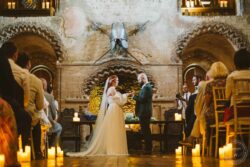 A romantic wedding altar in a rustic stone hall, featuring medieval-inspired design, candles, and a mounted suit of armour, capturing a special moment between the bride and groom during their celebration. Elegant wedding ceremony taking place in a historic stone chapel with medieval decor, illuminated by candlelight, with the bride and groom exchanging vows.