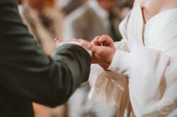 A bride and groom exchange rings at their wedding ceremony, captured in a detailed close-up showcasing love and commitment, with soft lighting and elegant attire. Close-up of a wedding ring being exchanged during a ceremony, focusing on hands and rings with blurred background and bridesmaid in white dress.