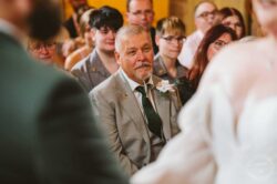 A wedding guest in a grey suit with a white boutonnière attentively watching the ceremony, amidst a diverse group of well-dressed attendees, in warm indoor lighting, capturing an emotional and memorable moment. Elegant wedding guest dressed in a grey suit with a white boutonnière, attentively listening at a wedding ceremony, surrounded by diverse guests in stylish attire, capturing a heartfelt moment.