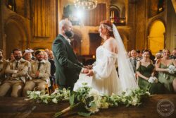 A bride and groom sharing their wedding vows inside a grand, historic hall, with guests smiling and witnessing the special moment, decorated with flowers and candlelit ambiance. Elegant wedding ceremony at a historic venue in Essex with a bride and groom exchanging vows, surrounded by joyful guests and warm ambient lighting.