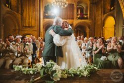 A romantic wedding moment with the bride and groom embracing at the altar, surrounded by happy guests clapping in a stunning historic hall in Essex & Suffolk. Elegant wedding couple sharing a kiss during ceremony in historic indoor venue with candlelit ambiance and joyful guests applauding in Essex & Suffolk.