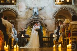 Romantic wedding at an Essex barn venue with a couple sharing a kiss during the ceremony, lit by candles and featuring medieval-inspired decor with mounted armour and stone arches. Elegant wedding ceremony in a historic Essex & Suffolk barn, capturing a heartfelt kiss between bride and groom surrounded by candlelit ambiance and rustic medieval decor.