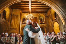 Beautiful wedding scene featuring the bride and groom embracing in a grand historic church with warm lighting and delighted guests celebrating their union. The venue showcases classic architecture with high wooden ceilings and intricate details, creating a timeless romantic setting. Elegant wedding couple sharing a kiss inside a historic Essex and Suffolk church, surrounded by joyful guests during their ceremony. Captured in a warm, romantic atmosphere with wooden beams and a chandelier.